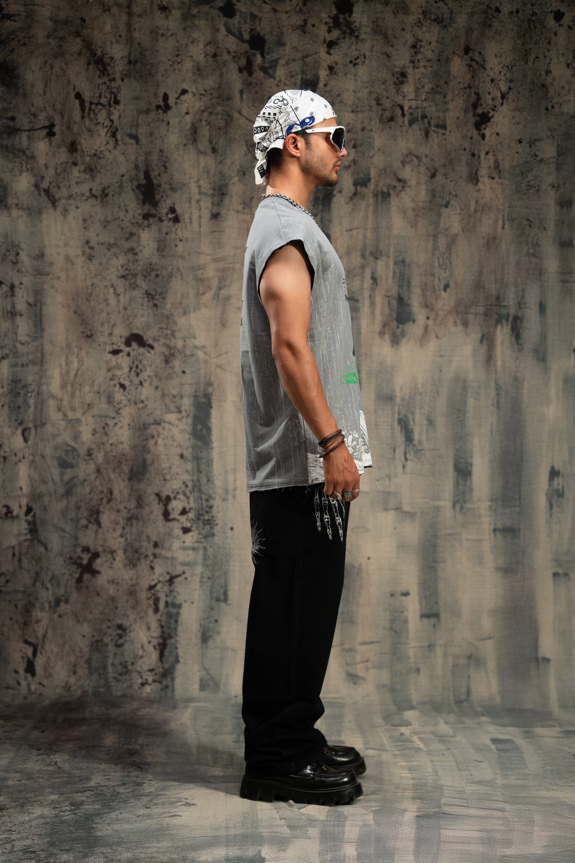Man wearing a sleeveless shirt and black pants standing against a textured wall.