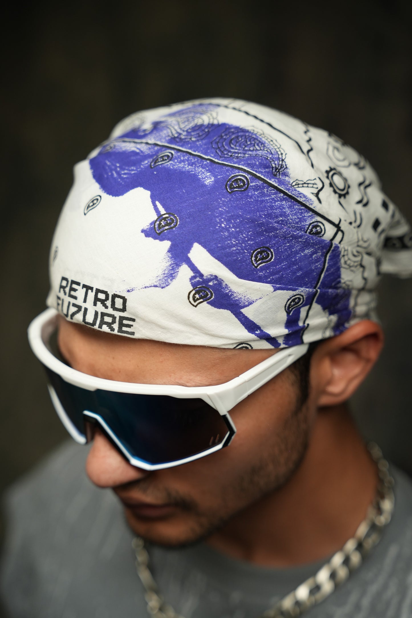 Person wearing a bandana with a graphic design and text, paired with sunglasses and a chain necklace.
