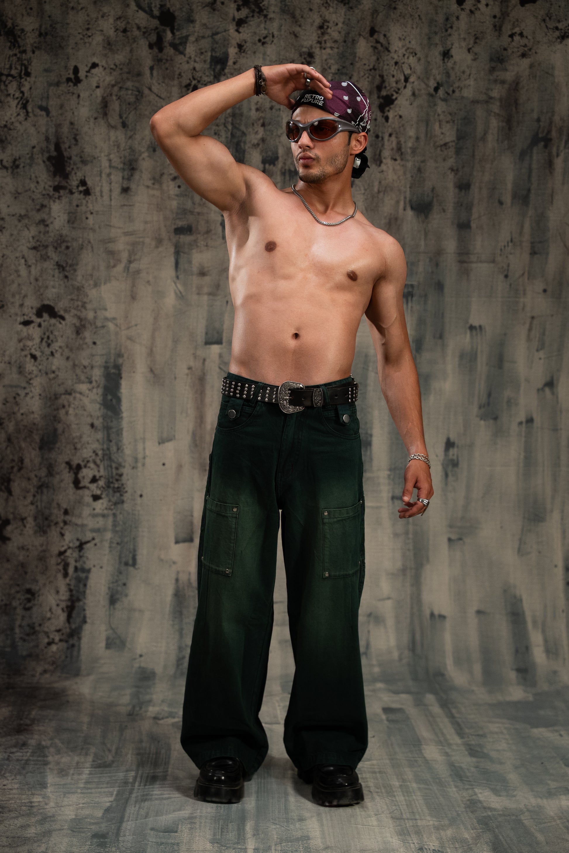 Man wearing dark green pants and a bandana against a textured wall.
