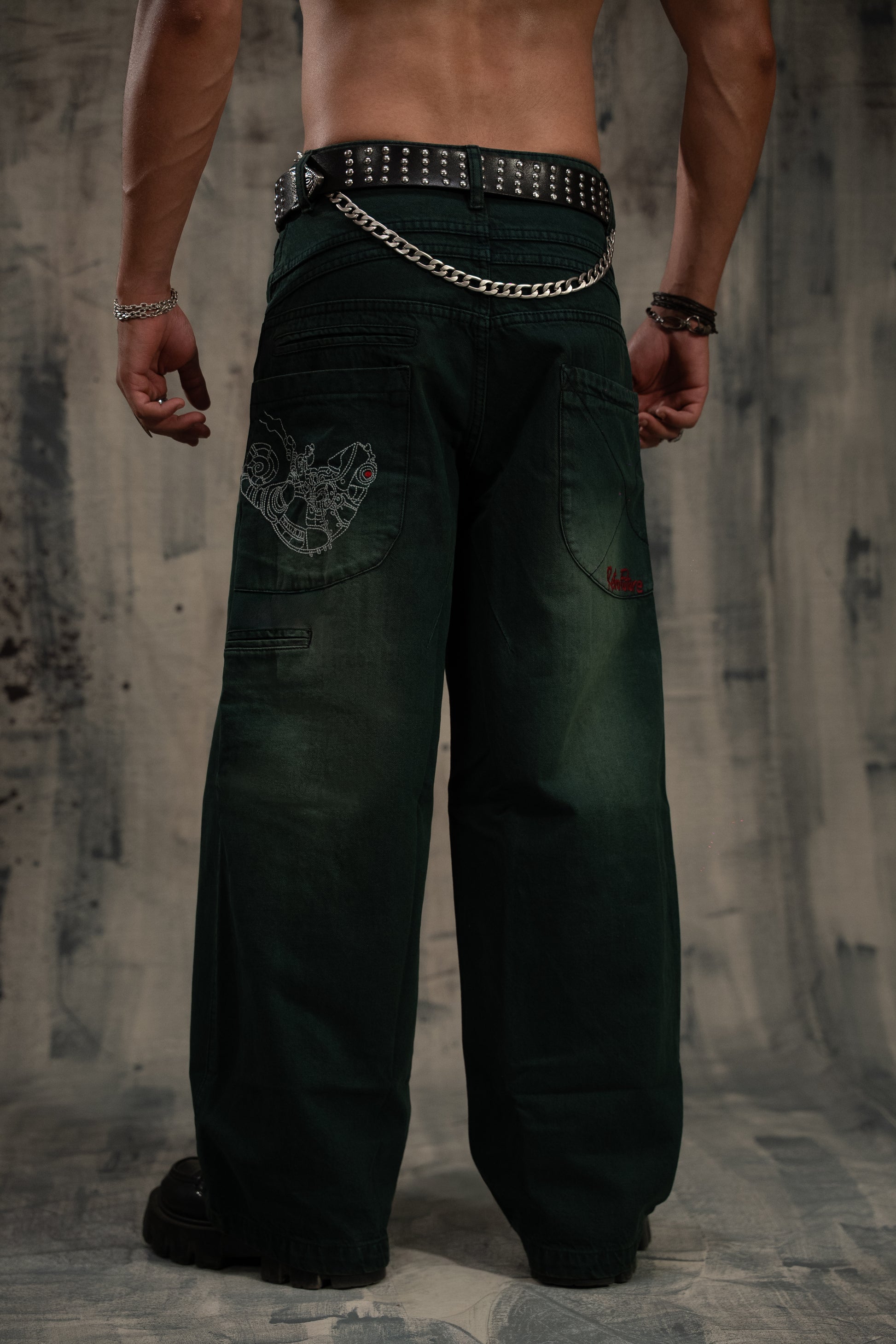 Person wearing dark green pants with chain detail and studded belt on a textured wall background