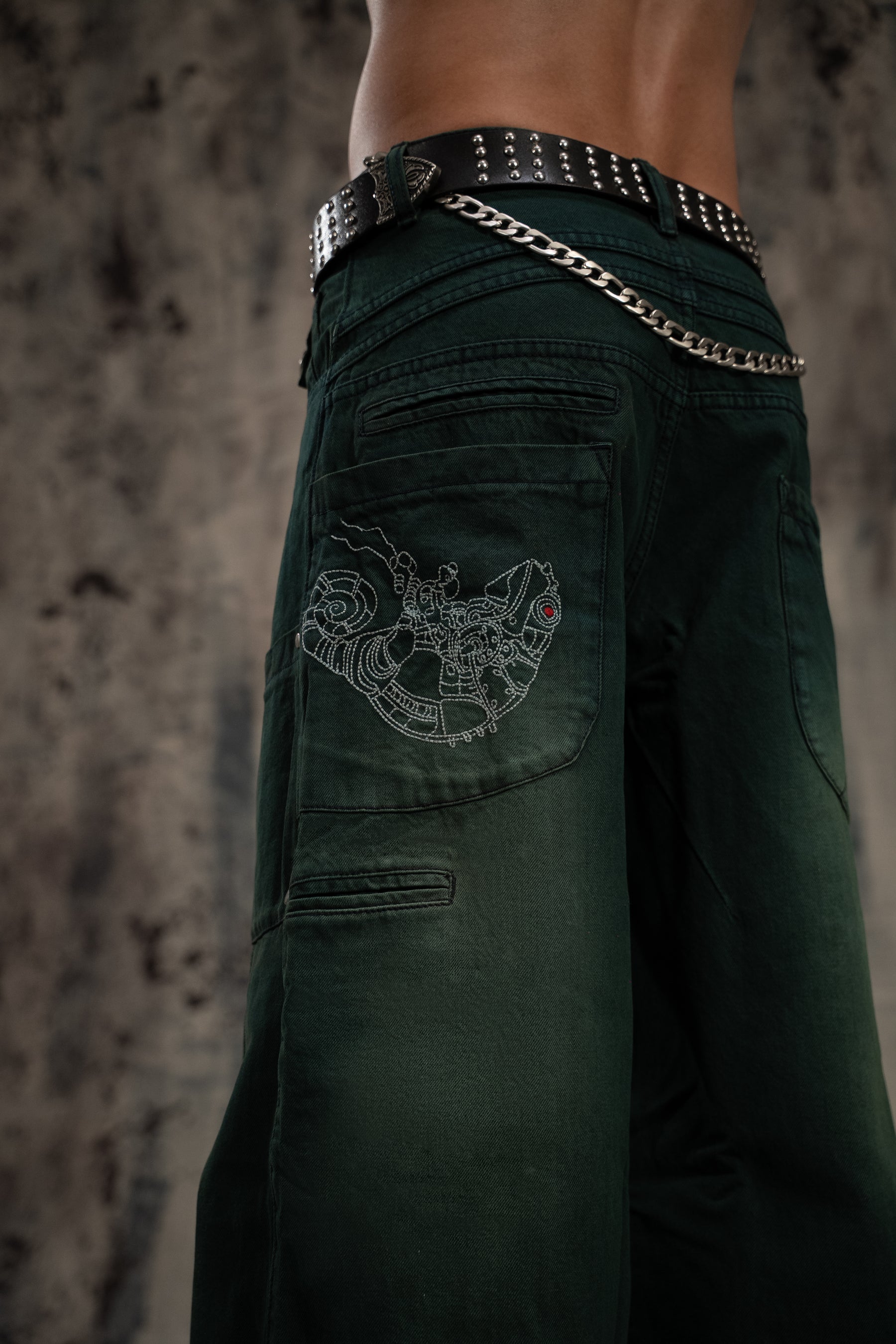 Dark green pants with a decorative belt and embroidered design on a textured background