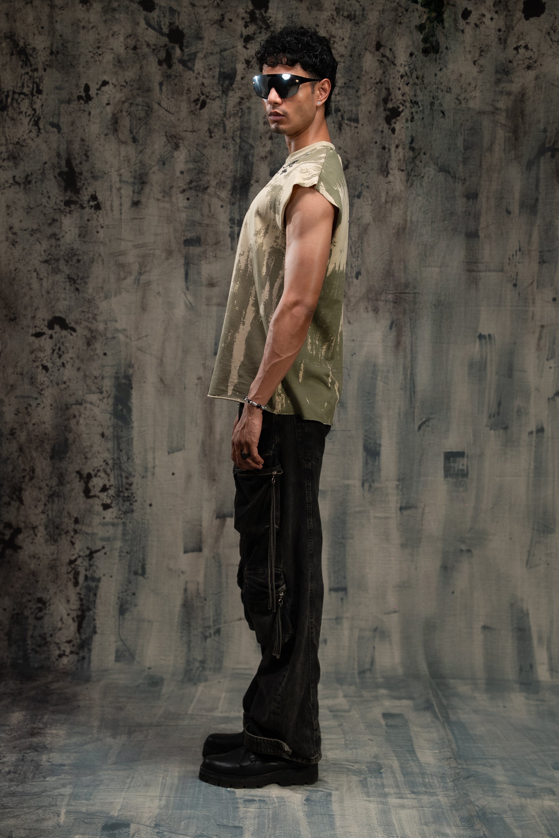 Man wearing a sleeveless green shirt and dark pants against a textured gray wall.