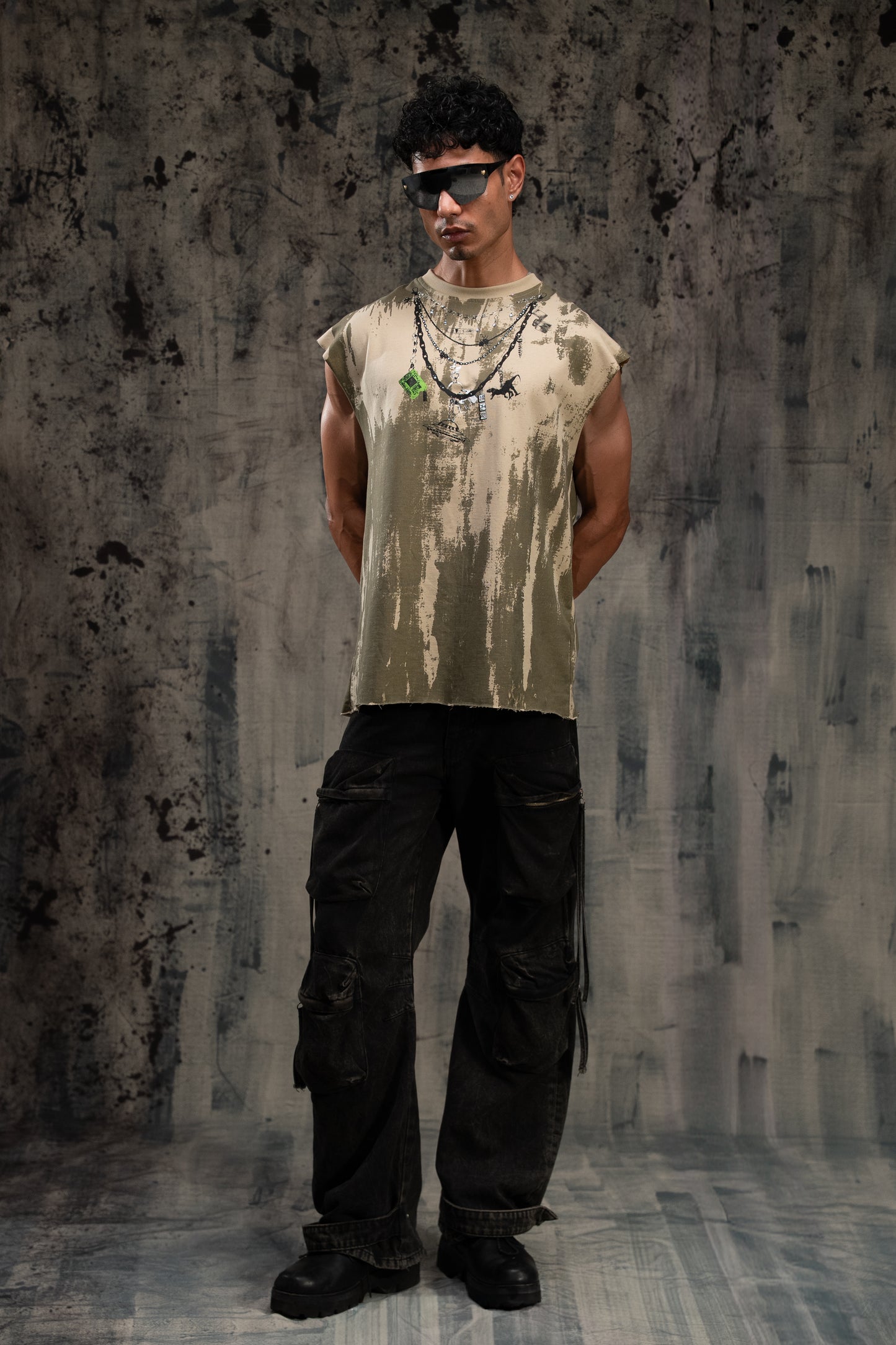 Person wearing a sleeveless distressed shirt and black pants against a textured wall.