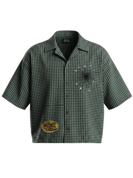 A short-sleeve button-up shirt in a green and black gingham pattern, featuring a broken glass graphic on the left chest with white spiderwebs and a circular yellow and black patch on the lower right side.